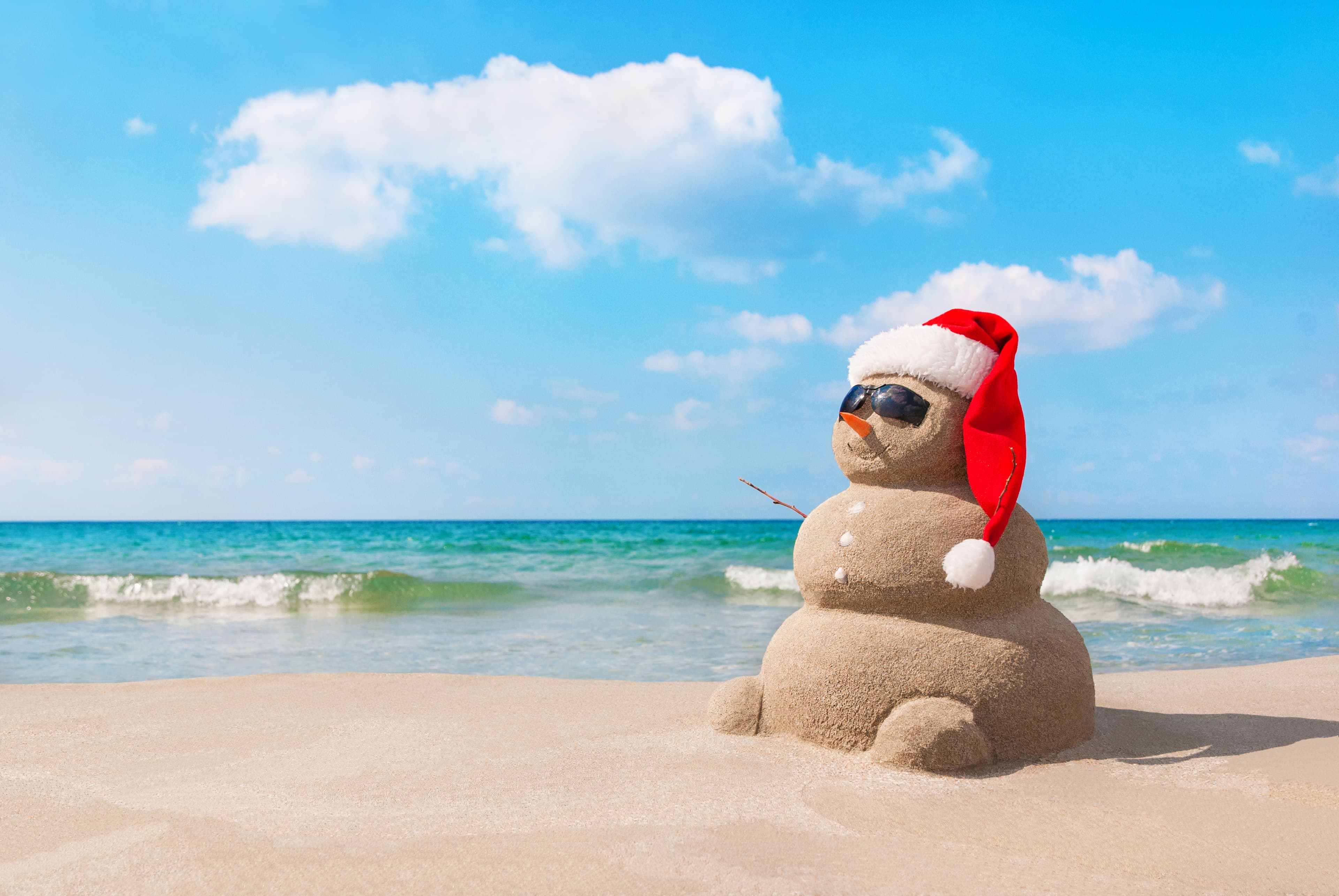 A sand snowman wearing a Santa hat and sunglasses on a sunny beach